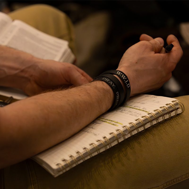 Close-up of a person's arms folded over an open notebook while taking notes during a Bible study or church service at Freedom Church in Murrysville, PA, wearing wristbands that read "PRAY" and "W.W.J.D."